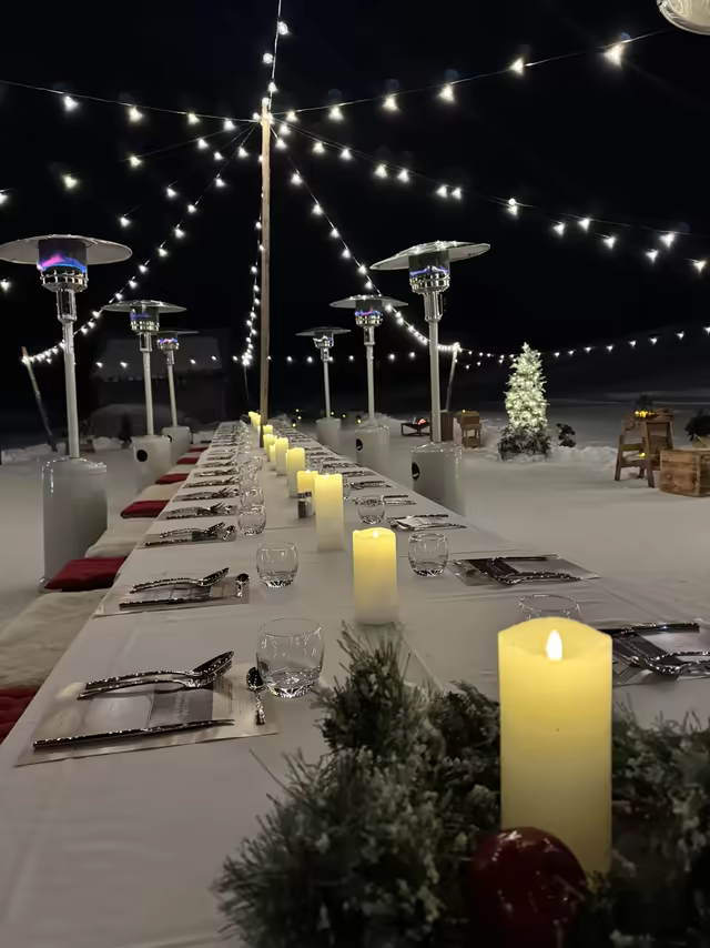 Long banquet table on the snow under string lights and candles, at night