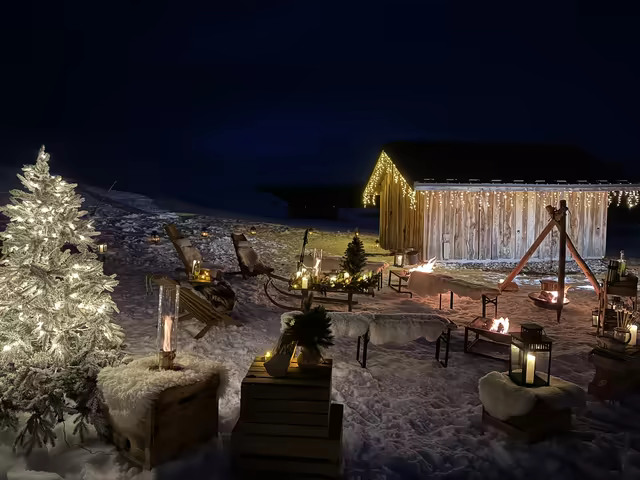 Wooden barn draped in fairy lights, with deck chairs and lanterns on the snow at blue hour
