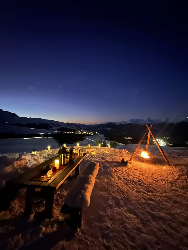 Candlelit table on a snowy slope at twilight, a campfire and the valley below