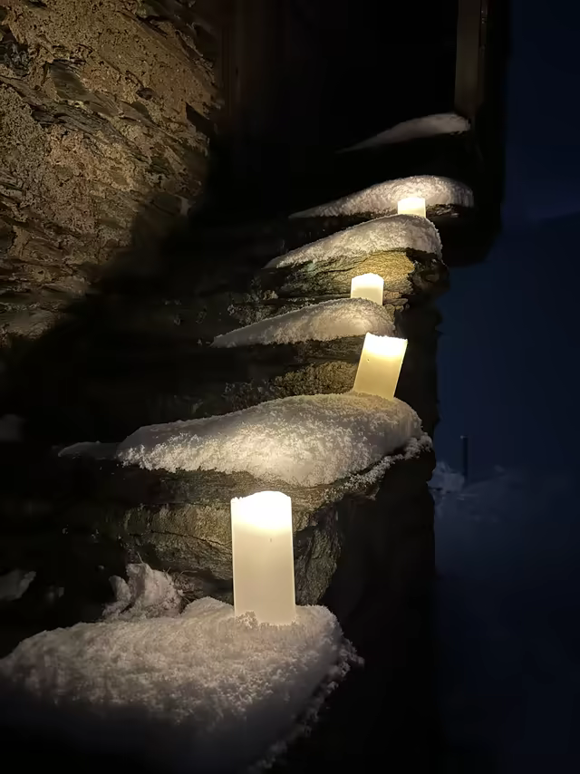 Candles lighting a snow-covered stone staircase along a chalet at dusk