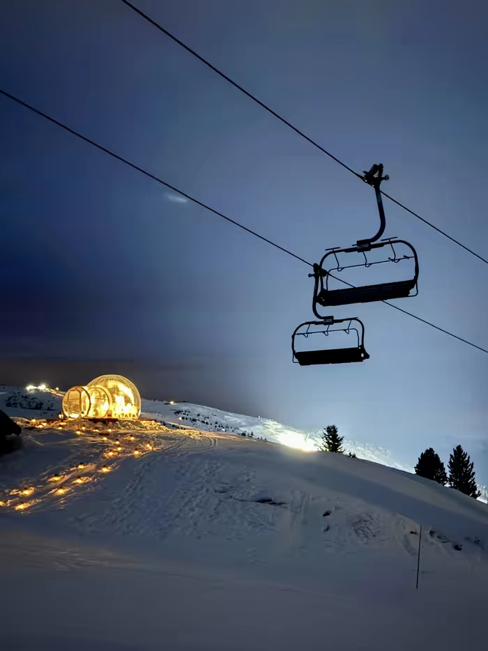 A chairlift passing above a glowing bubble and a path of lights on the snowy ridge at blue hour