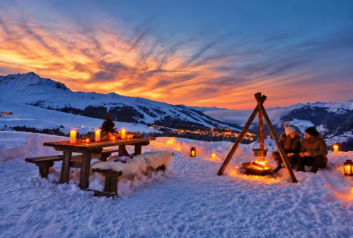 A couple by a fire under a tripod beside a candlelit table, facing the sunset over the peaks