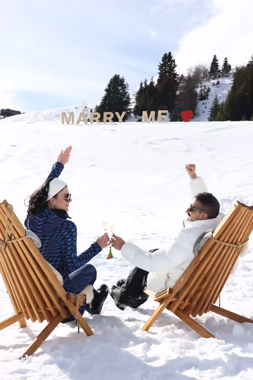 The couple raising their glasses from two wooden deck chairs, the Marry Me letters on the hill