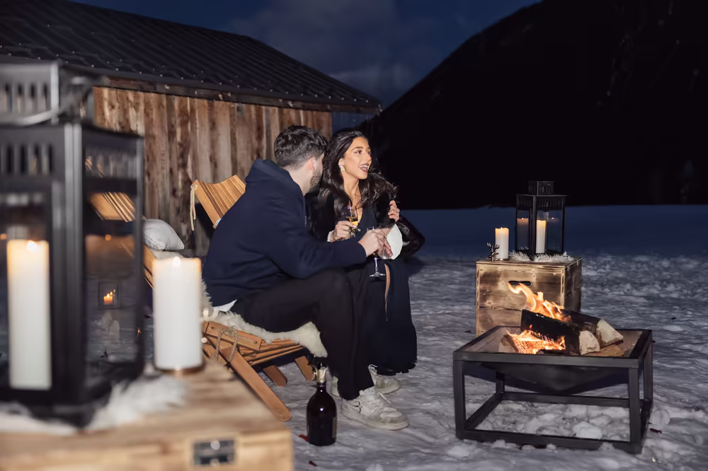 A couple sitting by a fire pit and lanterns against the chalet wall, at blue hour