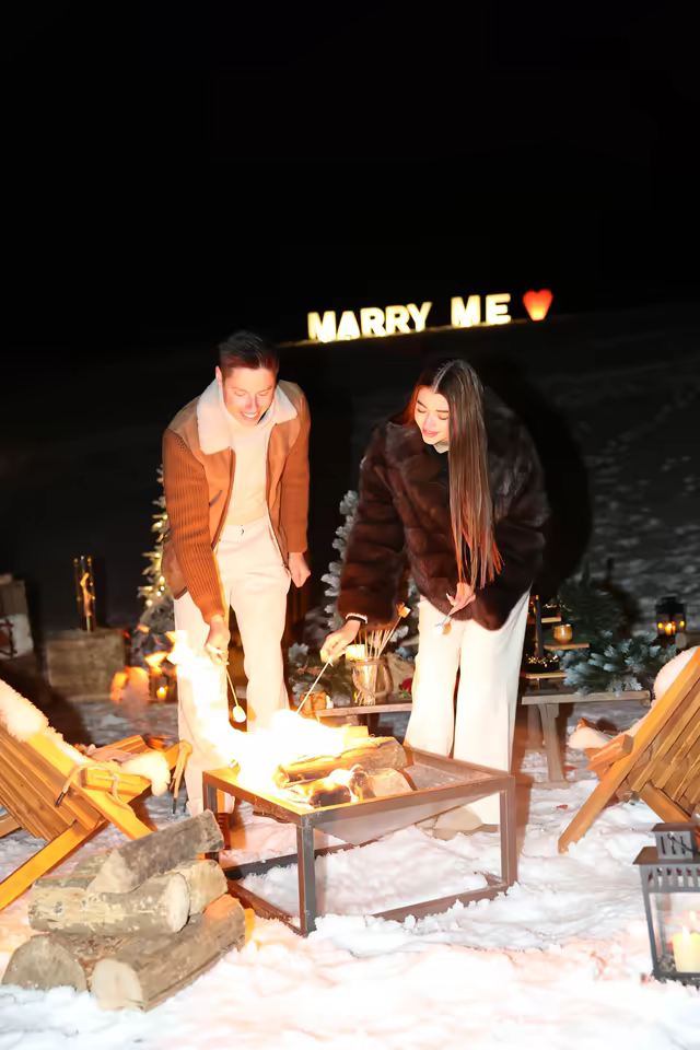 Couple at a fire table under an illuminated Marry Me sign, in the snow at night