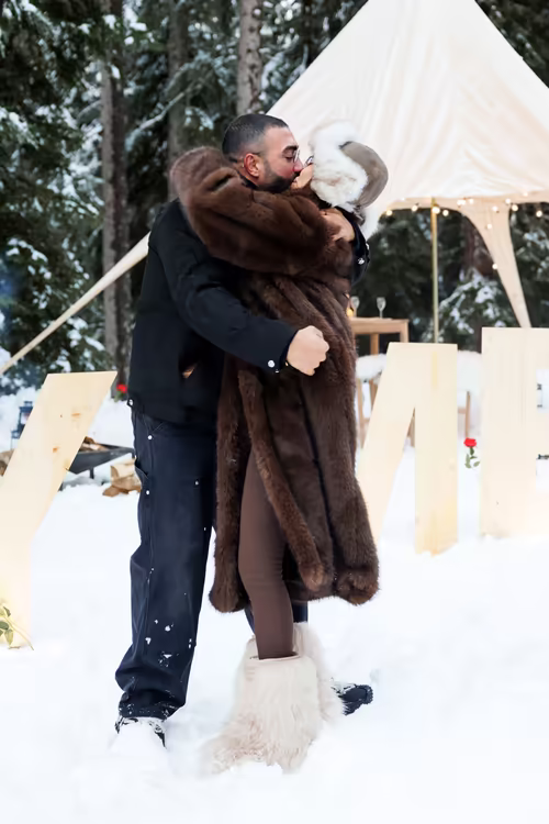 A couple kissing in the snow beside the wooden letters, in the forest clearing