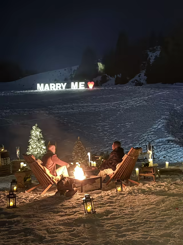 Marriage proposal by a fire pit, facing illuminated Marry Me letters in the snow