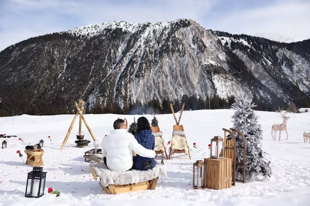 A couple seated on a sheepskin bench facing the mountain, lanterns and wooden reindeer around them
