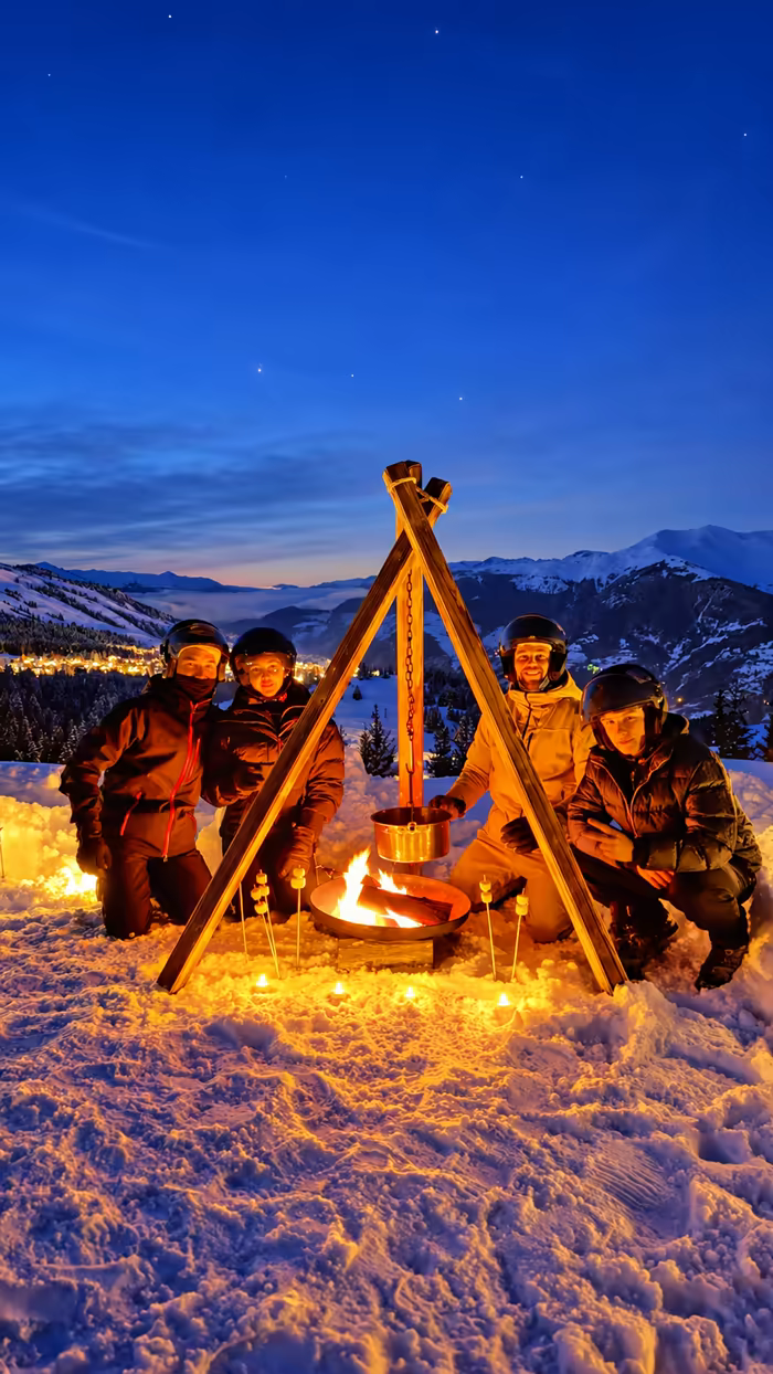 Four guests in helmets around a fire under a tripod at blue hour, above the valley