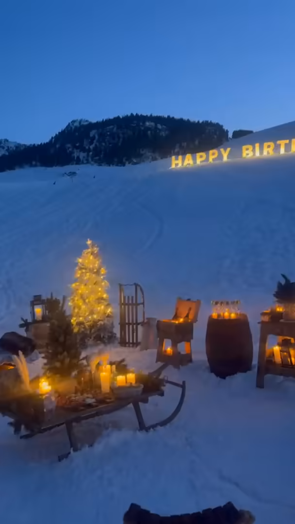 Lit Happy Birthday letters on the slope above a lit fir tree, a sledge and lanterns, at dusk