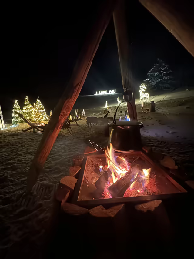 Illuminated I Love You sign by a campfire, among decorated fir trees in the snow