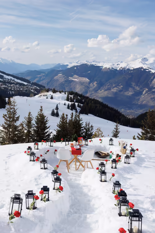 A path of lanterns and red roses leading across the snow to a table for two, high above the valley