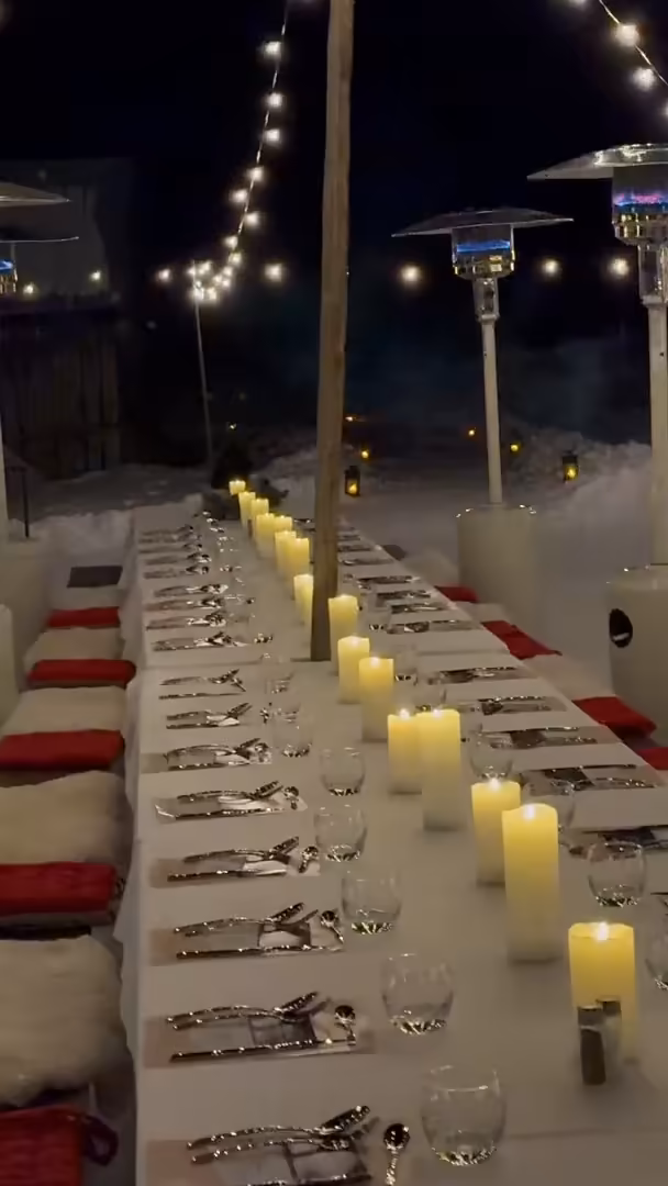 A long table set with candles under string lights and patio heaters, on the snow at night