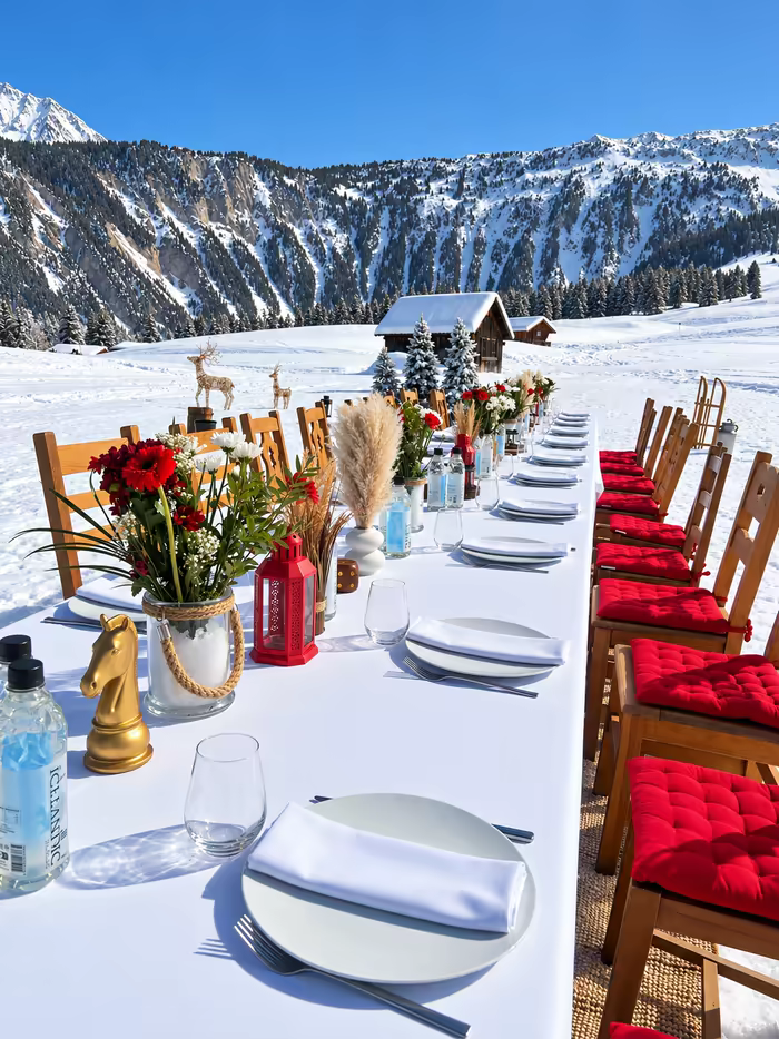 A long table laid in the snow, wooden chairs with red cushions, chalets and peaks in full sun