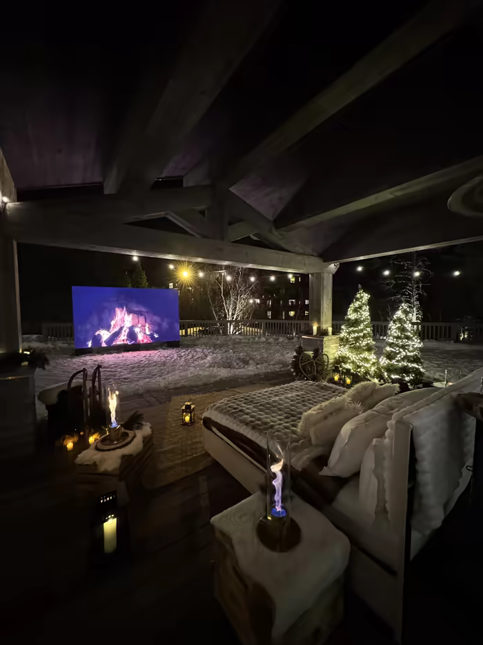 Snowy terrace lounge with a fireplace screen, a Christmas tree and flame lamps, at night