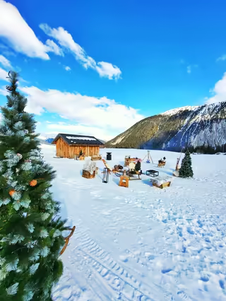 Wooden cabin, sleighs, fir trees and a campfire, surrounded by snowy mountains