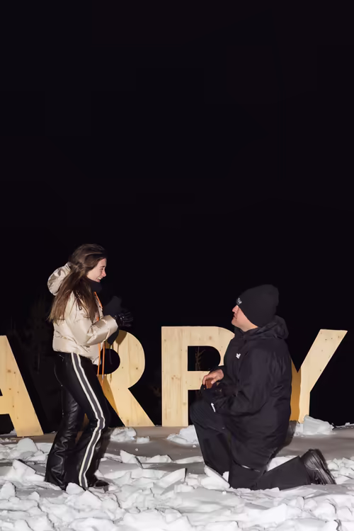 A man on one knee proposing in front of the Marry Me letters, at night