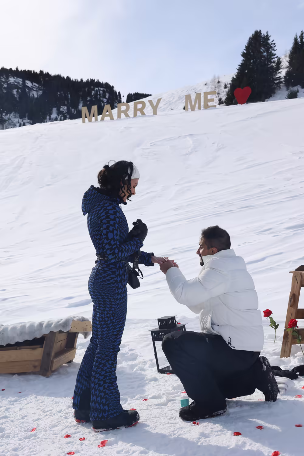 A man on one knee holding out the ring, the Marry Me letters on the slope behind