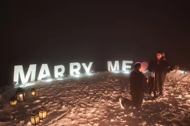Man on one knee before illuminated Marry Me letters and lanterns, on a snowy crest above Courchevel