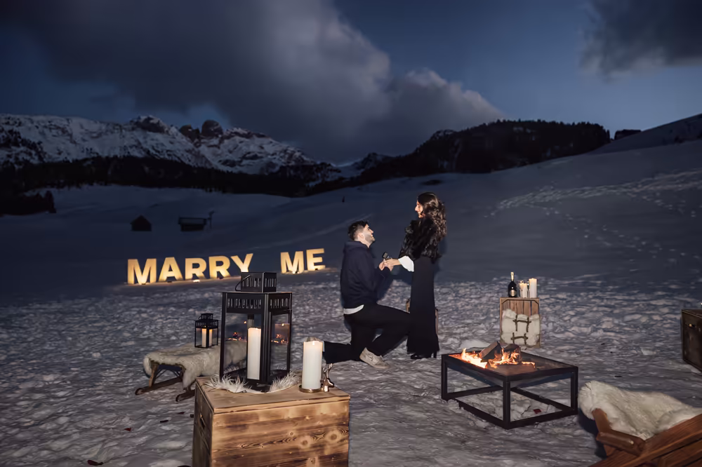 A proposal on one knee before the glowing Marry Me letters, the mountains in blue twilight