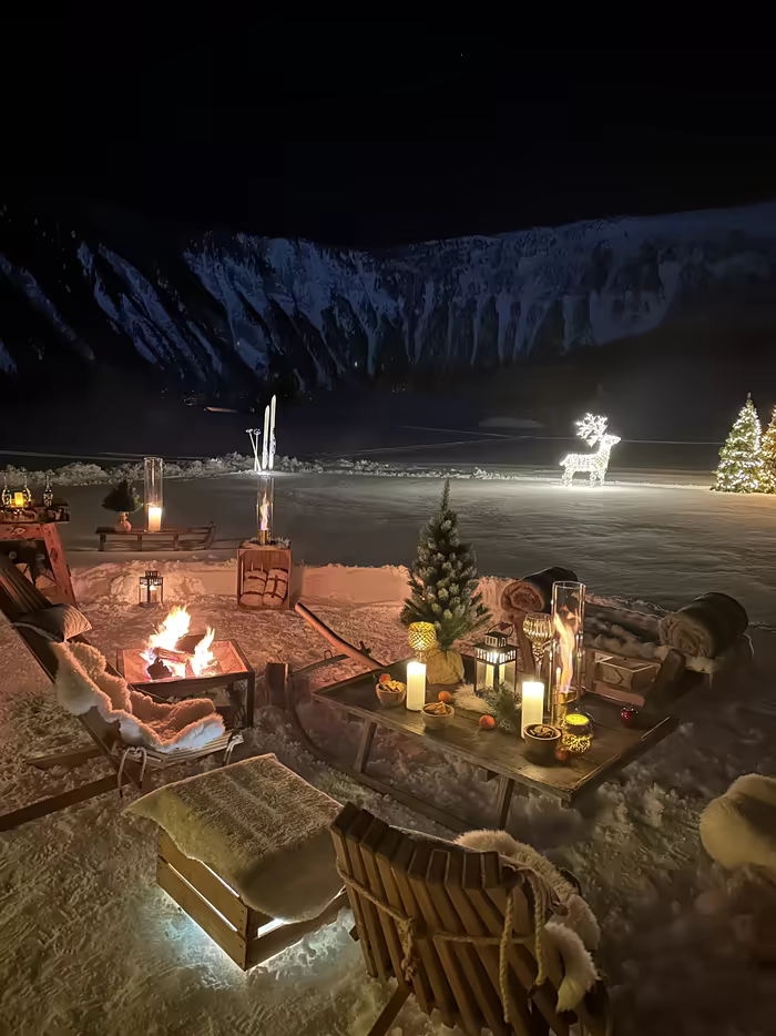Fire pit, candles and champagne on fresh snow, among lit reindeer and fir trees