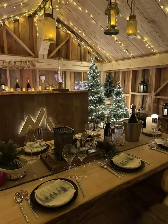 A table set in a wooden chalet under fairy lights, beside a Christmas tree and champagne