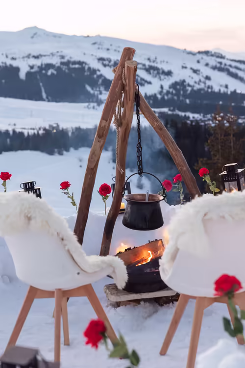 Two sheepskin-draped chairs around a fire under a cauldron, red roses planted in the snow