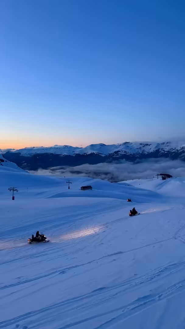 Snowmobiles with their headlights on crossing the slope at dusk, above a sea of clouds