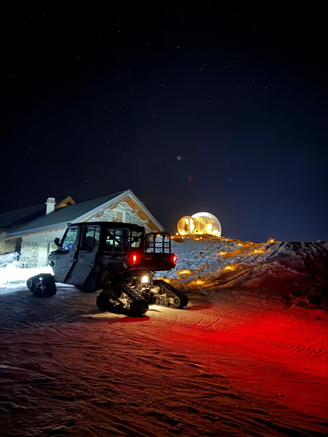 Tracked snow vehicle before a chalet at night, a lit bubble under the stars behind