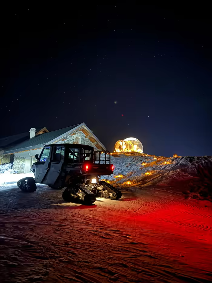 Tracked snow vehicle before a chalet at night, a lit bubble under the stars behind