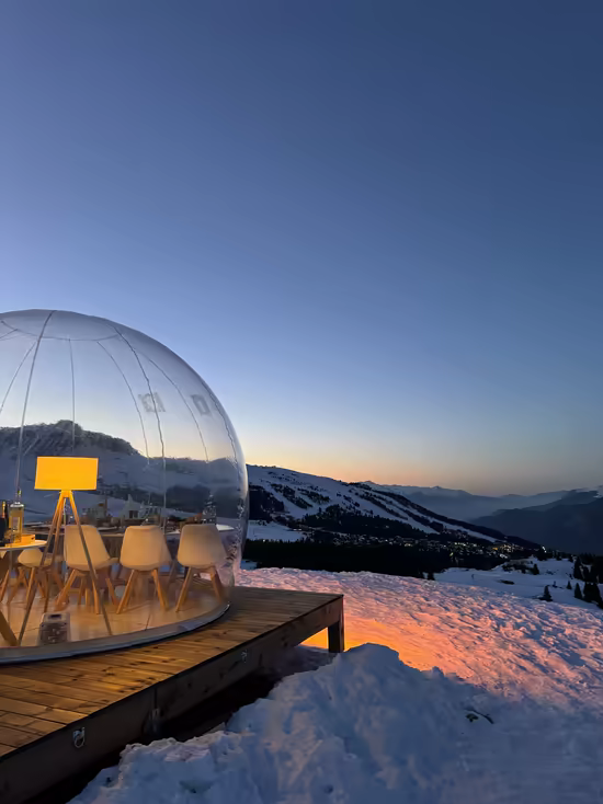 A transparent dining bubble on its wooden deck, the mountains glowing at sunset
